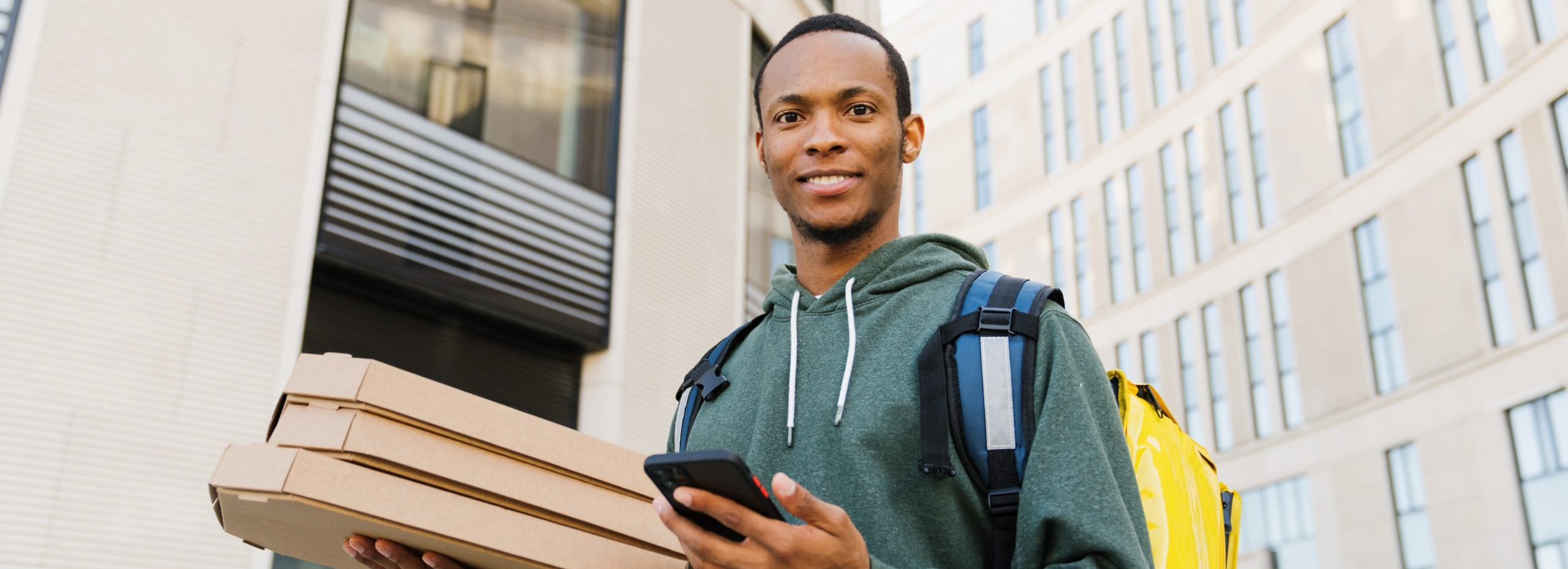 Pizza delivery. An African-American courier holds boxes outside and looks at the camera with a smile.